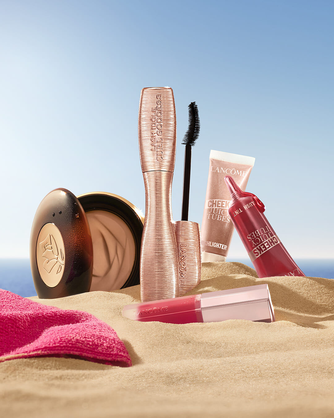 מוצרי איפור וטיפוח מבית לנקום (Lancome) מעוצבים על חול ים תחת שמיים בהירים, כולל מסקרה Lash Idole Curl Goddess בגוון זהב-ורוד, שפתוני Cheeks Juicy Tubes, ומייק-אפ קומפקט לצד מגבת ורודה.