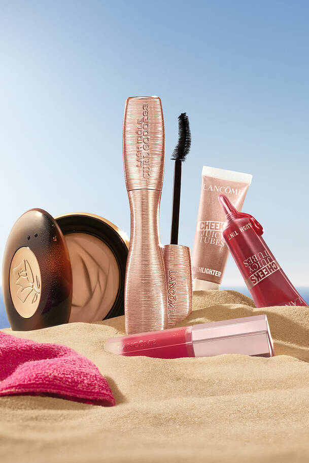 מוצרי איפור וטיפוח מבית לנקום (Lancome) מעוצבים על חול ים תחת שמיים בהירים, כולל מסקרה Lash Idole Curl Goddess בגוון זהב-ורוד, שפתוני Cheeks Juicy Tubes, ומייק-אפ קומפקט לצד מגבת ורודה.